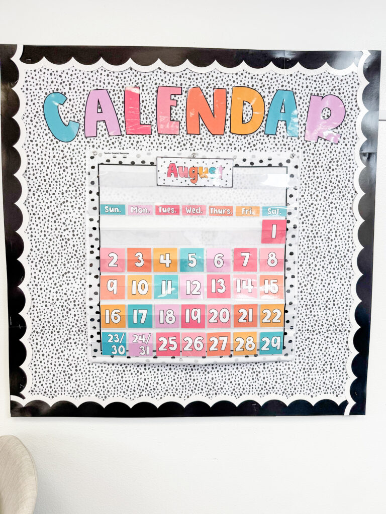 This image shows a calendar display on a bulletin board. The calendar display is within a pocket chart that has a white background and a border of black polka dots surrounding the entire calendar. 