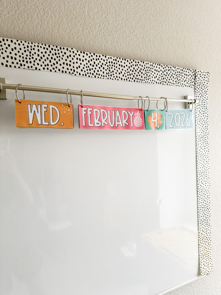 This image shows a close-up of a flip calendar attached to a whiteboard. The cards show the day of the week, month, date, and year. They are brightly colored, feel like paradise and have small designs in the background.