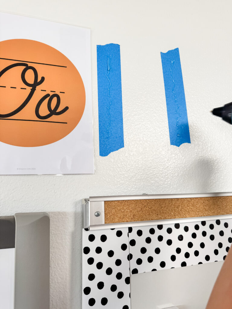 This image shows a hot glue gun applying hot glue on top of two strips of blue painter's tape.