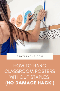 This image shows a teacher hanging a poster above a whiteboard without using staples. The text at the bottom of the image reads How to Hang Classroom Posters Without Staples (No Damage Hack!)