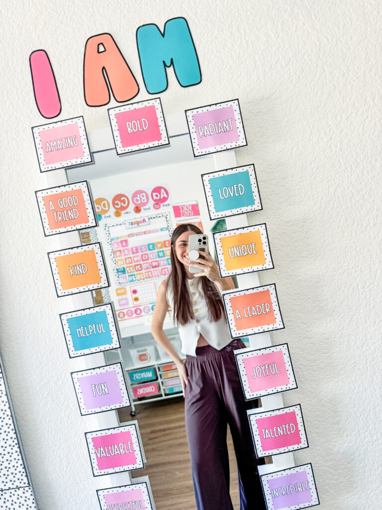 This image shows an affirmation station. There is a white mirror on a wall with pastel squares all around with different "I am" positive affirmation statements.