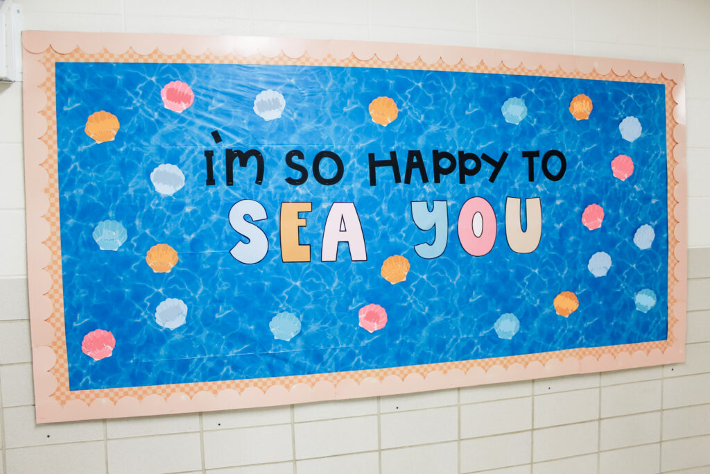 This image shows a bulletin board outside of a 2nd grade classroom that reads I'm so happy to sea you. It's decorated to look like the sea with colorful seashells all over. 