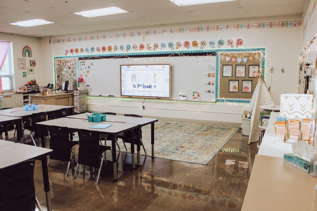 This image shows a front of a 2nd grade classroom with a rug in front of the whiteboard. There are lots of twinkle lights around the bulletin boards and lamps used to create a cozy feel. 