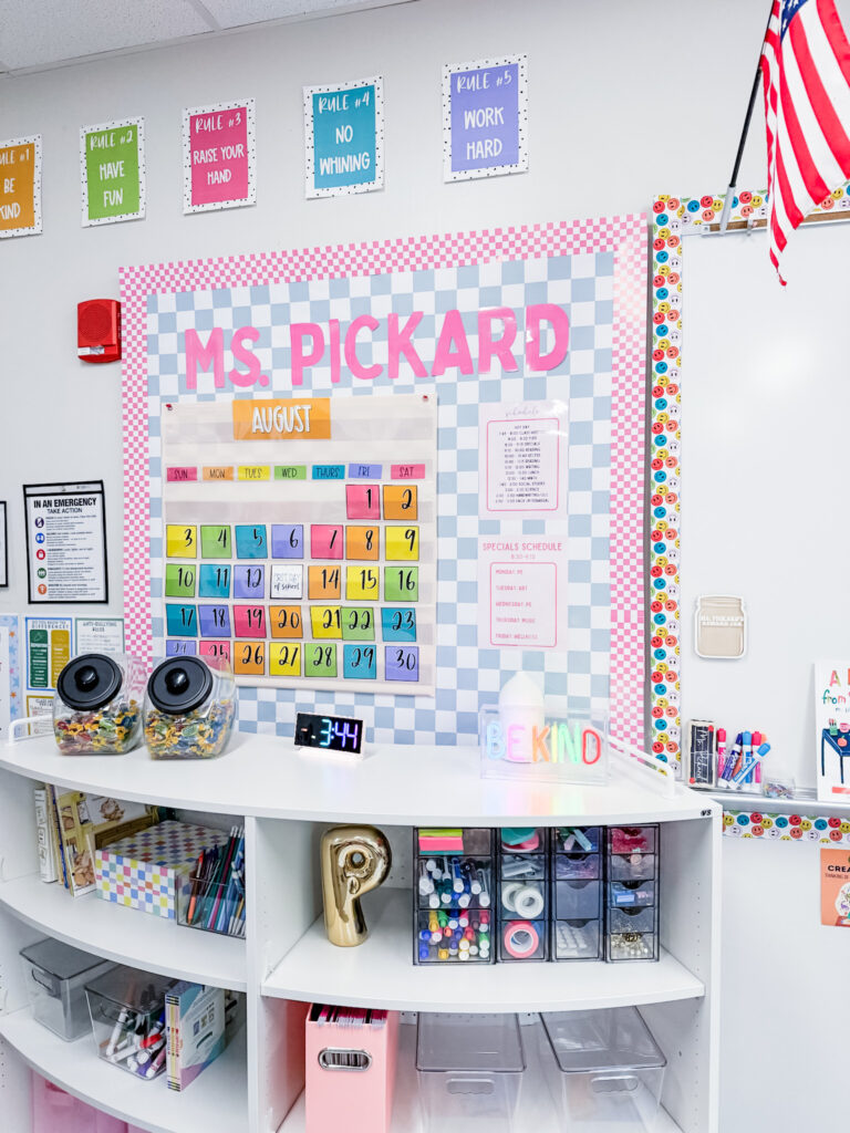 This image shows a curved shelf at the front of a classroom filled with markers, teacher supplies, and a calendar on the bulletin board.