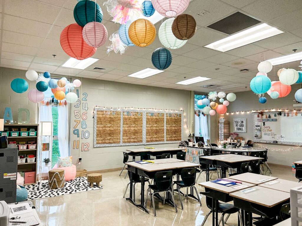 This image shows a whimsical 3rd grade classroom with paper lanterns hanging from the ceiling, desks in groups of 4, a cozy reading nook in the corner, and twinkle lights surrounding the entire classroom.
