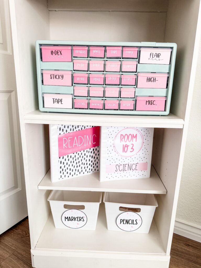 This image shows a bookcase with a teacher toolbox that has pink labels on it showing what goes in each section. On the lower shelves are two three ring binders and two white bins for markers and pencils. 
