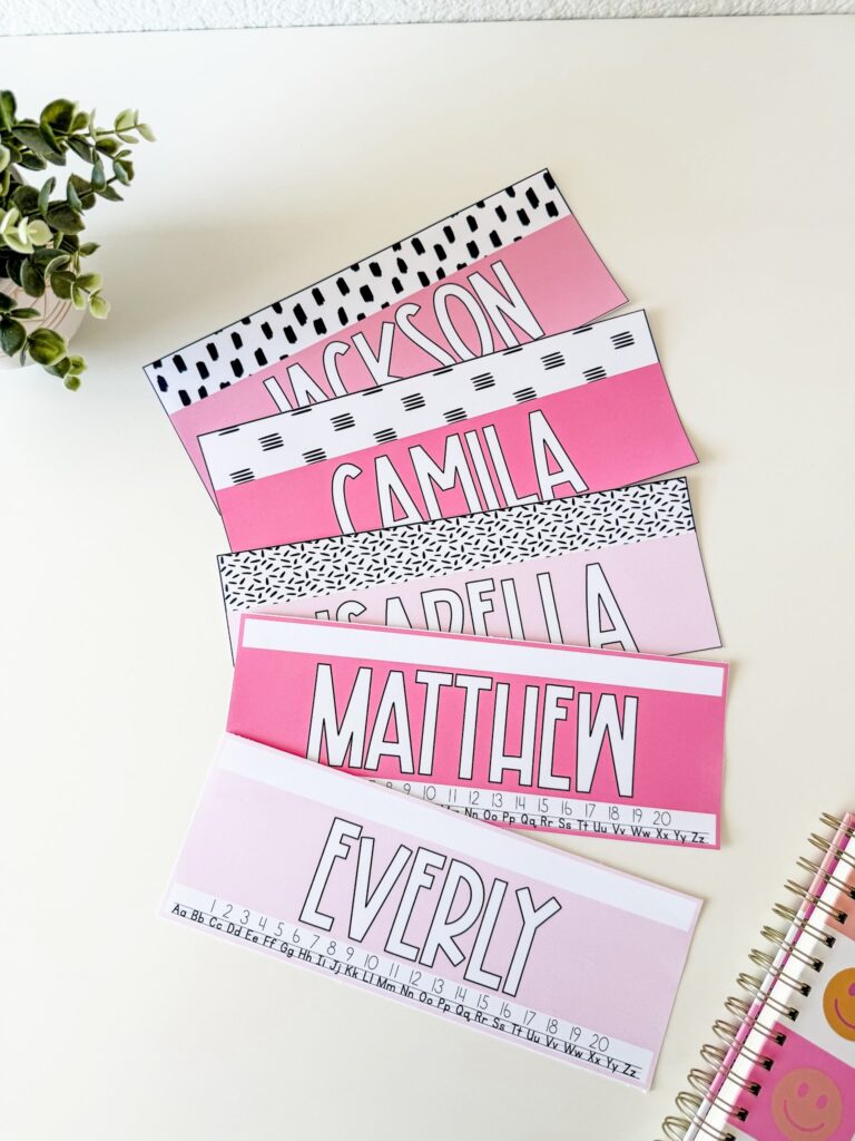 This image shows five different nametag templates included in the simple + pink classroom decor bundle. Some nametags just have a solid pink background with  a decorated pattern along the top. Other nametags have a number line and the alphabet written underneath for a reference. 