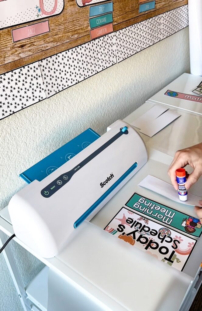This image shows a teacher using a glue stick to glue down small pieces she wants to laminate from her classroom decor before sending them through the laminator.