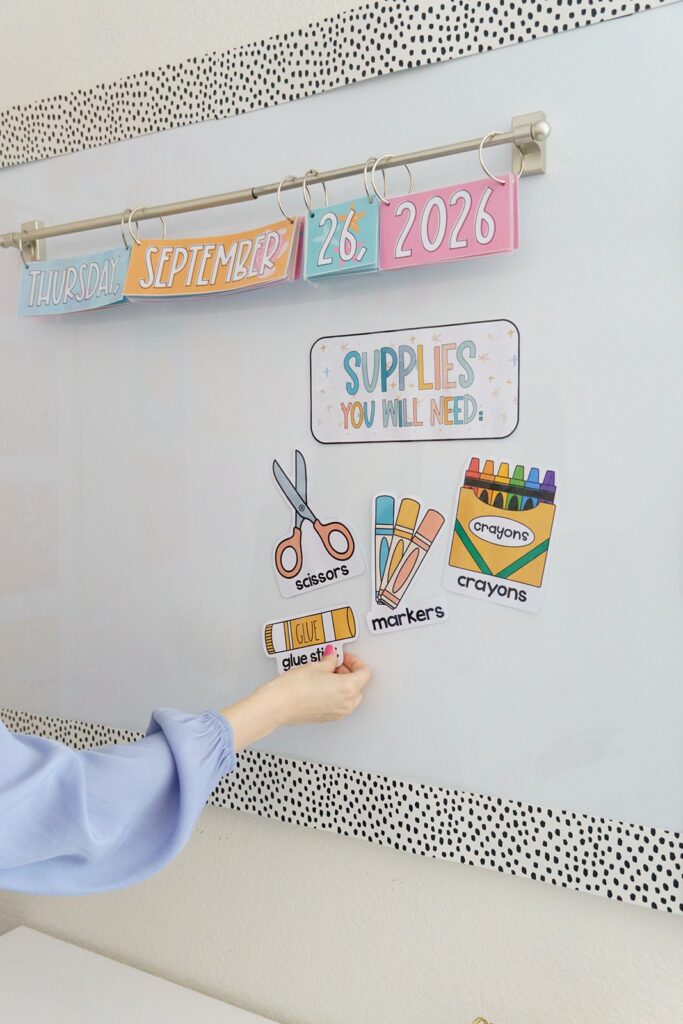 This image shows a teacher adding cutout images of different supplies to her whiteboard based on what her students need for the upcoming lesson. There is a banner that reads "Supplies You Will Need". Underneath that banner there are the following images: scissors, markers, crayons, and glue sticks. 