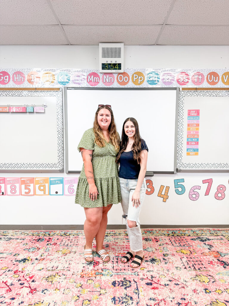 This image shows two young ladies standing in front of a whiteboard of a 4th grade math classroom.