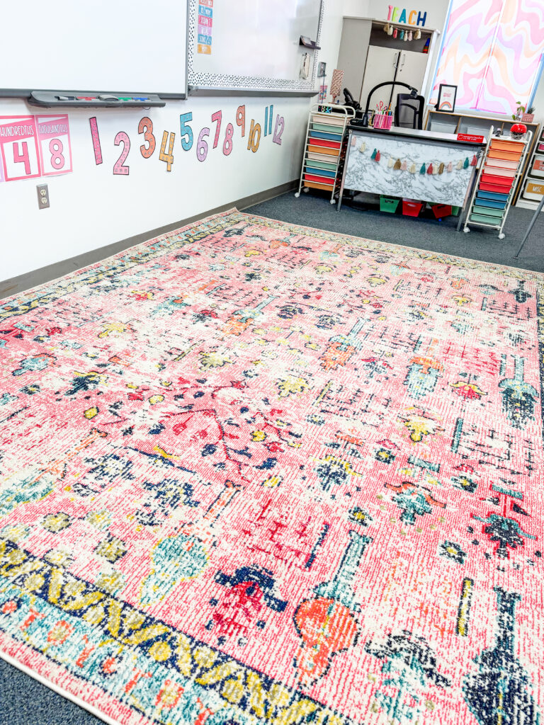 This image shows a brightly colored, mostly pink background, rug in the front of a classroom. It's the perfect fit for the pastel classroom decor decorating this space.