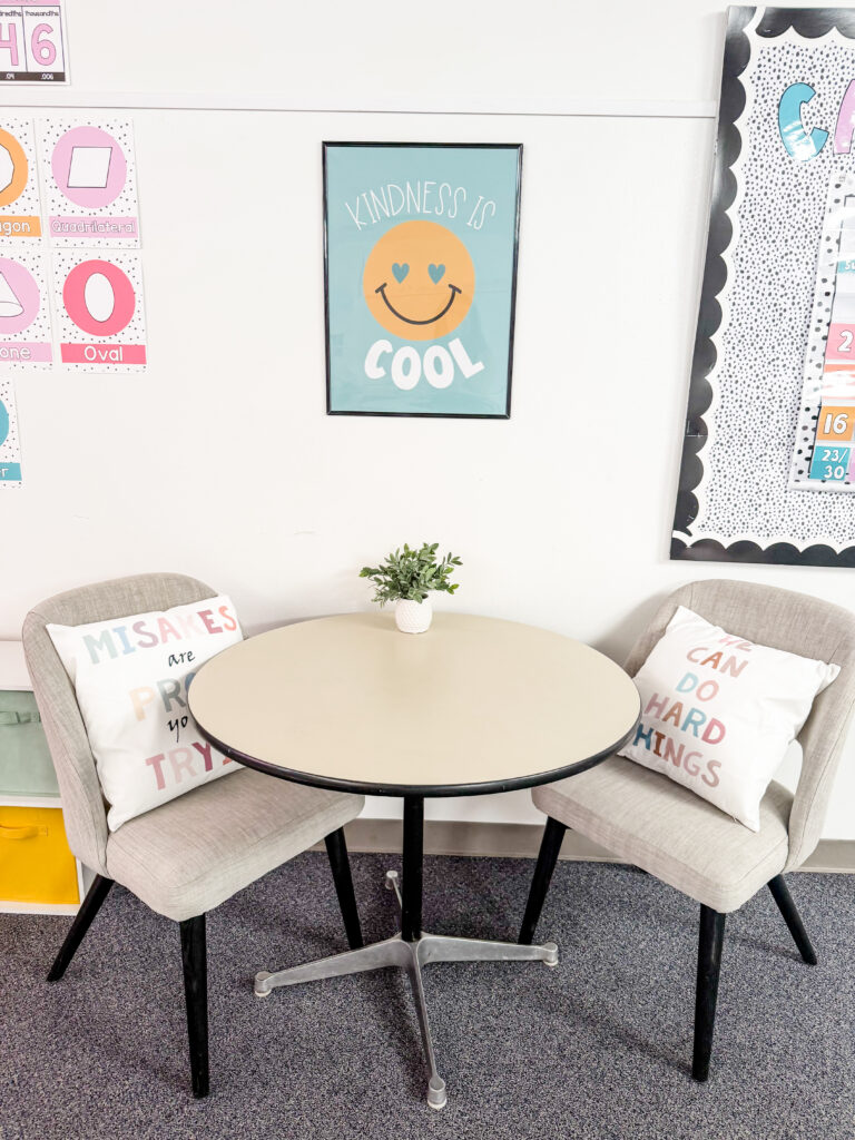 This image shows a close-up of a small, circular table in a classroom with two gray chairs around it with throw pillows and a poster on the wall that reads "Kindness is Cool".