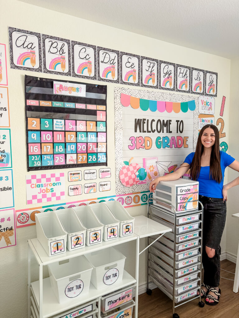 This image shows a teacher standing by her classroom decked out in pastel composition decor.