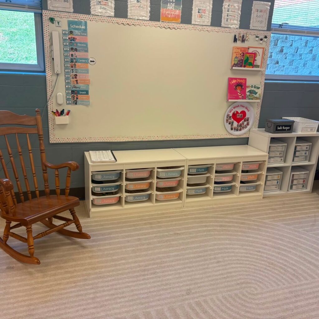 This image shows a close-up of the front of the classroom. There are visual schedule cards posted on the whiteboard, as well as shelves with picture books. Underneath the whiteboard are more storage areas with bins for math and literacy time.