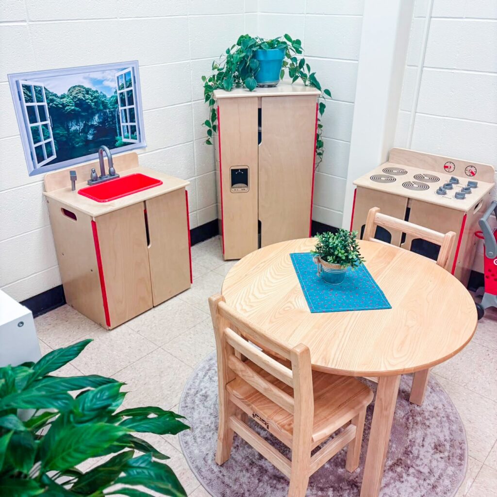 This image shows a play area in a pk classroom. There is a wooden sink, fridge, stove, and table with two chairs.