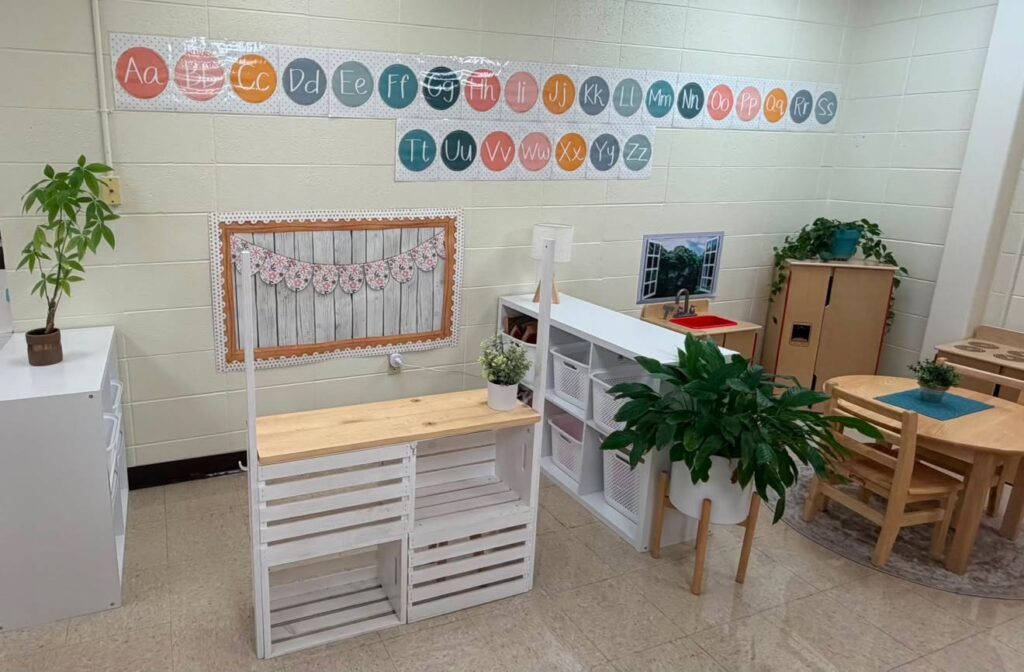 This image shows a wooden stand in a pre-k classroom. By the wooden stand, there are other cube organizers with baskets holding different supplies. Along the wall are alphabet posters. 